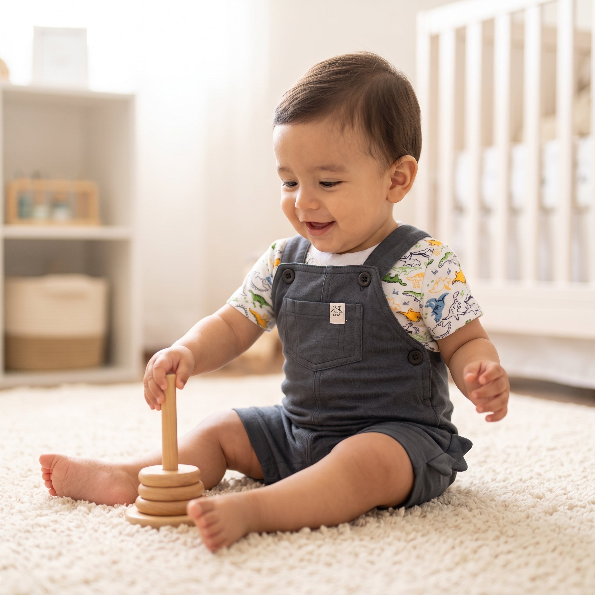 lifestyle: A new photorealistic photograph, not a modification of any input image. The scene is a bright, cozy nursery room with soft, diffused natural light pouring in from a window, illuminating a plush cream-colored rug. A cheerful baby boy of Latino/Hispanic ethnicity, 9-12 months old, is sitting playfully on the rug. He is wearing the product, with the t-shirt visible layered underneath the romper, and the dinosaur print clearly visible on the t-shirt. His posture subtly highlights the dinosaur tail d…