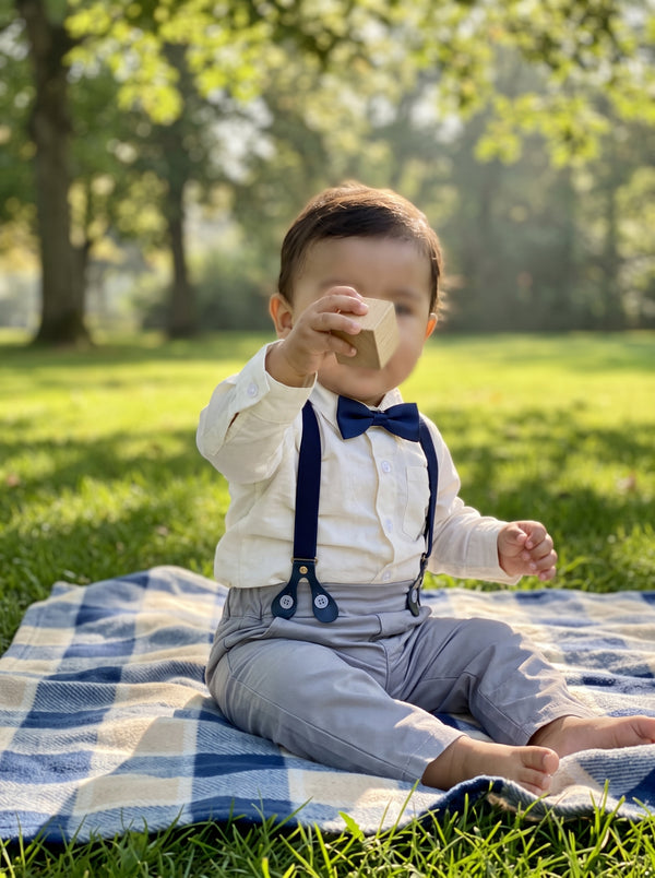 lifestyle: A NEW photorealistic photograph composed from scratch, depicting a vibrant outdoor park setting with lush green grass and a soft, checkered picnic blanket spread out. Sunlight filters through overhead trees, creating dappled patterns on the ground. A Latino/Hispanic baby boy, 10-12 months old, with a playful demeanor, is sitting upright on the picnic blanket wearing the product. His hand is reaching for a simple baby toy (e.g., a wooden block or soft fabric ball) that partially obscures his face…